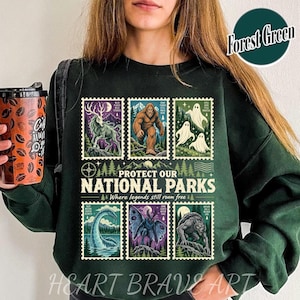 May include: A dark green long-sleeve sweatshirt featuring a graphic print of six postage stamp-style illustrations depicting mythical creatures and the text Protect Our National Parks Where legends still roam free in cream lettering.