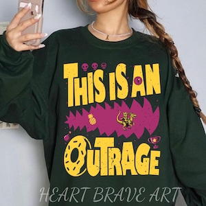 Puede incluir: Sudadera verde oscuro con el texto amarillo "THIS IS AN OUTRAGE", junto con un gr&aacute;fico de un dinosaurio montando una criatura. La sudadera tambi&eacute;n presenta una rosquilla, una pi&ntilde;a y un trofeo. Las palabras "HEART BRAVE ART" est&aacute;n en la parte inferior.