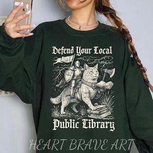 Può includere: Felpa verde scuro con la scritta "Defend Your Local Public Library". La grafica presenta un cavaliere che cavalca un gatto, che tiene una bandiera e un'ascia, con una pila di libri. Le parole "Heart Brave Art" sono in basso.