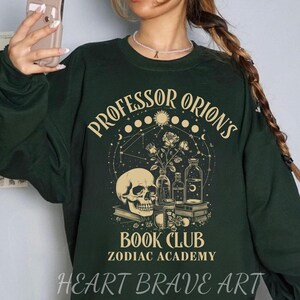 May include: Green sweatshirt with a graphic design featuring a skull, flowers, and bottles. The text "Professor Orion's Book Club Zodiac Academy" is printed on the sweatshirt.