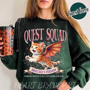May include: A forest green sweatshirt with the text "QUEST SQUAD" in pink lettering. The sweatshirt features a cartoon illustration of an orange cat with dragon wings and the text "BROCCOLI'S VERSION" in a banner. The text "NOBODY WANTS IT BUT IT'S GOOD FOR YOU" is printed below the banner.