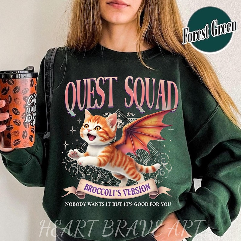 May include: A forest green sweatshirt with the text "QUEST SQUAD" in pink lettering. The sweatshirt features a cartoon illustration of an orange cat with dragon wings and the text "BROCCOLI'S VERSION" in a banner. The text "NOBODY WANTS IT BUT IT'S GOOD FOR YOU" is printed below the banner.