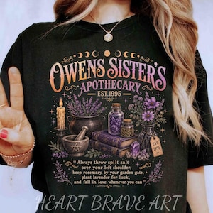 Könnte beinhalten: Ein schwarzes T-Shirt mit einem Grafikdruck, der den Text "OWENS SISTER'S APOTHECARY EST. 1995" sowie das Zitat "Always throw spilt salt over your left shoulder, keep rosemary by your garden gate, plant lavender for luck, and fall in love whenever you can." zeigt.
