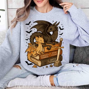 May include: Light gray sweatshirt featuring a dragon design. The graphic includes two dragons, one brown and one gold, perched on a stack of books with a sword. The books have the titles "Rebecca Yarros", "Iron Flame", and "Onyx Storm".