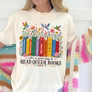 May include: Cream-colored t-shirt featuring a colorful graphic of books, flowers, and birds. The text "It's a good day to READ QUEER BOOKS" is displayed below the books. The design includes book titles like "Heartstopper" and "One Last Stop".