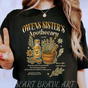 May include: A black t-shirt featuring a graphic print with the text Owens Sister's Apothecary est. 1995. The design includes a glass bottle, a mortar and pestle with herbs, cinnamon sticks, and additional text that reads There's a little witch in all of us.