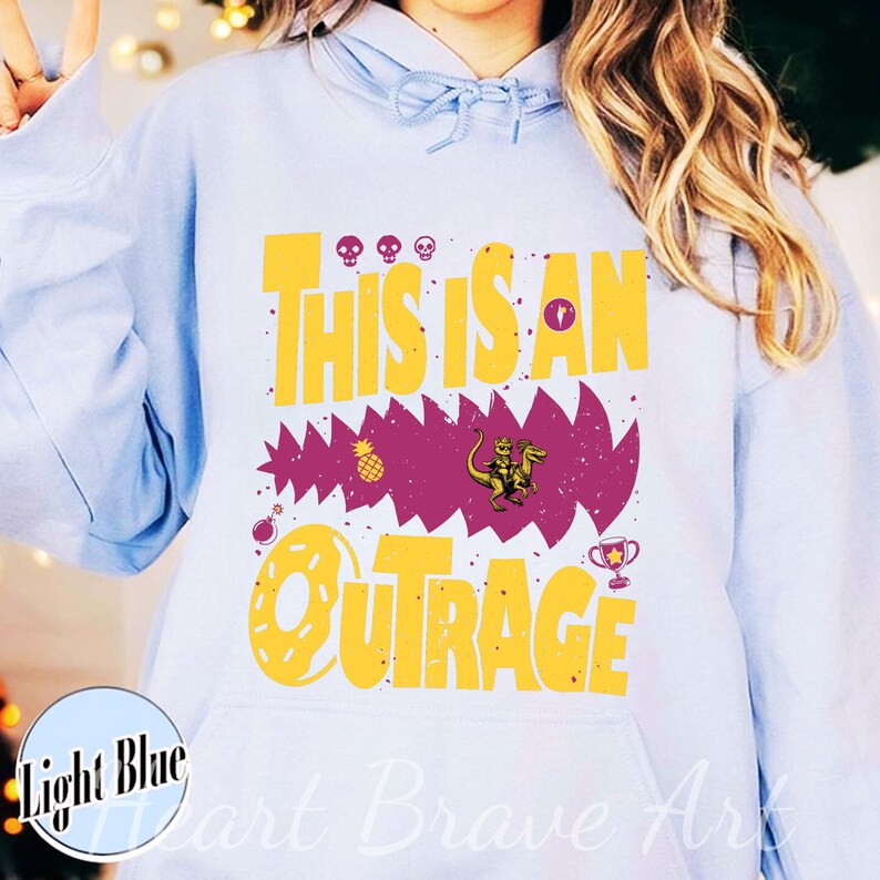 Puede incluir: Sudadera con capucha azul claro con el texto "THIS IS AN OUTRAGE" en amarillo. El dise&ntilde;o incluye una forma dentada p&uacute;rpura con un dinosaurio montando una criatura, una pi&ntilde;a y otros gr&aacute;ficos.