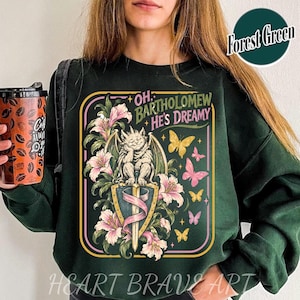 May include: A forest green crewneck sweatshirt featuring a graphic print of a gargoyle perched on a shield with a sword, surrounded by pink lilies and yellow and pink butterflies. The text reads, Oh, Bartholomeow He's Dreamy, in a stylized font.