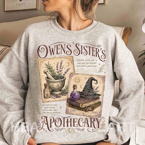 May include: A light gray crewneck sweatshirt featuring a graphic print with the text "OWENS SISTER'S APOTHECARY". The design includes illustrations of a mortar and pestle with lavender, a purple potion bottle, a witch hat, and stacked books.