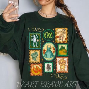 Könnte beinhalten: Dunkelgrünes Sweatshirt mit einer Collage von Illustrationen im Briefmarkenstil zum Thema Der Zauberer von Oz. Die Briefmarken zeigen den Zinnmann, die Vogelscheuche, Dorothys Kleid und den Löwen. Der Text "OZ" und "HEART BRAVE ART" sind ebenfalls sichtbar.