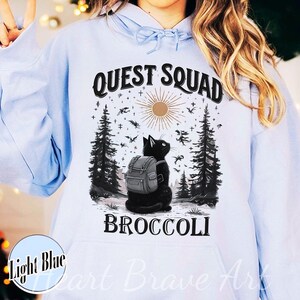 May include: Light blue hoodie with a graphic featuring a black cat wearing a backpack, set in a forest scene with the text "QUEST SQUAD BROCCOLI". The design includes a sun, trees, and flying creatures.
