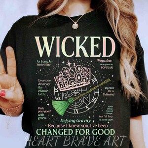 Op de afbeelding: Zwart T-shirt met het woord "WICKED" in lichtroze letters. Het shirt heeft een roze kroon, een groene bezem en zinnen uit de musical. De tekst "HEART BRAVE ART" staat onderaan.