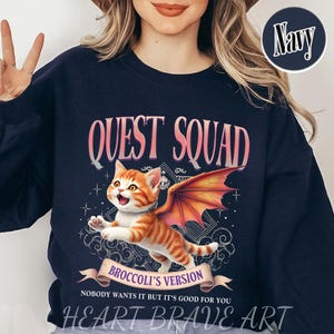 May include: A navy blue sweatshirt with a graphic of a cartoon orange cat with wings. The text "QUEST SQUAD" is above the cat and "BROCCOLI'S VERSION" is below the cat. The text "NOBODY WANTS IT BUT IT'S GOOD FOR YOU" is below the graphic. The text "HEART BRAVE ART" is at the bottom of the sweatshirt.