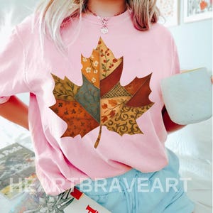 Puede incluir: Sudadera rosa claro con un diseño de hoja de arce grande y colorida. La hoja está compuesta por varios parches estampados en tonos naranja, marrón y verde. El texto "HEARTBRAVEART" es visible en la parte inferior.