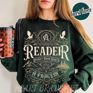 Op de afbeelding: Groen sweatshirt met een grafisch ontwerp met een lezer met een boek, draken en de tekst "A Reader Without Their Book Is A Tragedy".