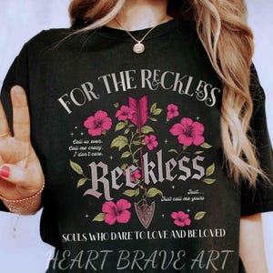 Puede incluir: Camiseta negra con un diseño gráfico que presenta flores rosas, una flecha y el texto "FOR THE RECKLESS", "Reckless", "Just call me yours" y "SOULS WHO DARE TO LOVE AND BE LOVED". La parte inferior de la camiseta tiene el texto "HEART BRAVE ART".