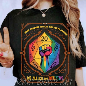 May include: Black t-shirt featuring a colorful graphic of a twenty-sided die with a raised fist and the text "When Someone Attacks One Party Member, We All Roll For Initiative." The design includes rainbow colors and decorative elements.