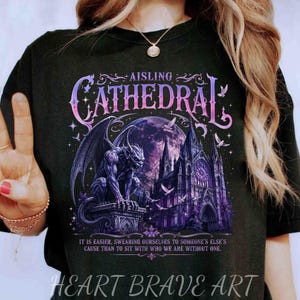 May include: A black t-shirt featuring a graphic print of a gargoyle perched on a stone pedestal in front of a gothic cathedral and a large purple moon. The text reads AISLING CATHEDRAL and includes a quote about swearing at the bottom.