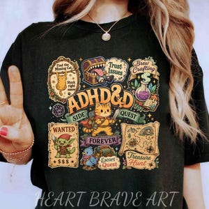 May include: A black t-shirt featuring a colorful graphic print with the text ADHD&D. The design includes a central orange cat adventurer surrounded by various fantasy-themed illustrations, including a treasure chest, a map, and a wanted poster.
