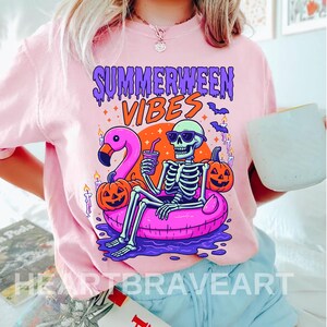 Könnte beinhalten: Hellrosa T-Shirt mit einem farbenfrohen Grafikdesign. Das Design zeigt ein Skelett mit Sonnenbrille auf einem rosa Schwimmring, mit einem Flamingo und Kürbissen. Der Text "SUMMERWEEN VIBES" befindet sich über dem Skelett.