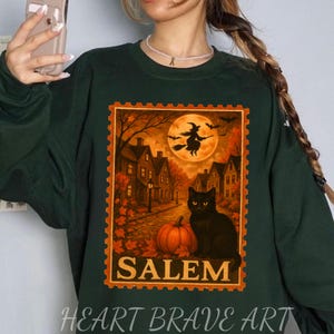 May include: Dark green sweatshirt featuring a Halloween-themed graphic. The graphic includes a black cat, pumpkin, witch on a broom, and the word "Salem" in orange. The design is framed like a postage stamp. The text "HEART BRAVE ART" is at the bottom.
