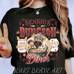 May include: Black t-shirt with a graphic featuring a cartoon Viking chef stirring a pot of soup. Text includes "Senshi's Dungeon Diner," "Fresh Monsters Daily," and "Open 24/7." The shirt also has the text "Heart Brave Art."