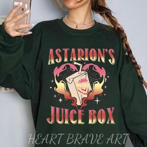 以下が含まれることがあります： 炎と「Astarion's Juice Box」というテキストが赤、オレンジ、黄色のグラデーションで書かれたジュースボックスのグラフィックプリントが施された濃い緑色のスウェットシャツ。スウェットシャツの下部にはグレーで「HEART BRAVE ART」というテキストがプリントされています。