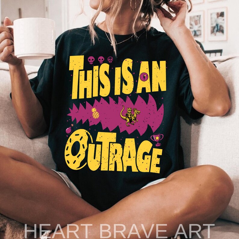 Puede incluir: Camiseta negra con el texto amarillo "THIS IS AN OUTRAGE", con un dise&ntilde;o morado dentado con un dinosaurio, una pi&ntilde;a y un trofeo. La camiseta es de corte holgado.