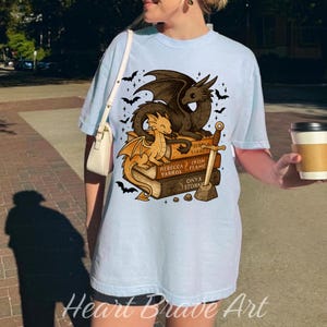May include: Light blue t-shirt featuring a dragon graphic. The design includes two dragons, a sword, and books with the titles "Rebecca Yarros", "Iron Flame", and "Onyx Storm". The shirt also has the text "Heart Brave Art".
