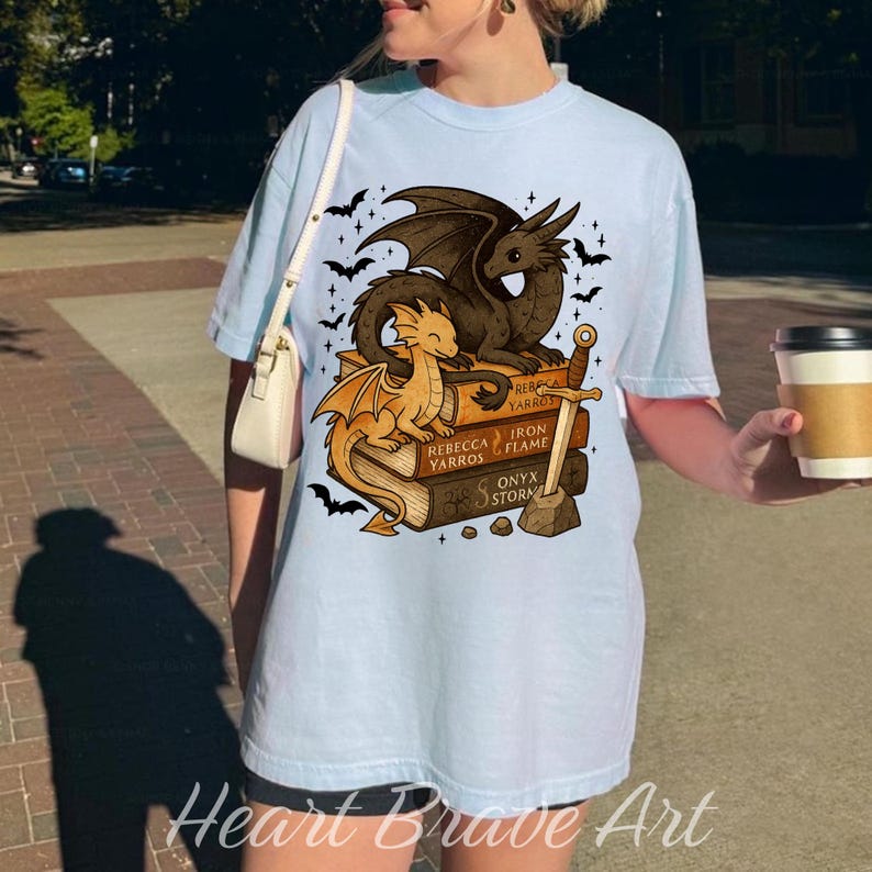 May include: Light blue t-shirt featuring a dragon graphic. The design includes two dragons, a sword, and books with the titles "Rebecca Yarros", "Iron Flame", and "Onyx Storm". The shirt also has the text "Heart Brave Art".