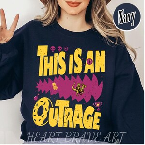 Puede incluir: Sudadera azul marino con el texto amarillo "THIS IS AN OUTRAGE", con un gr&aacute;fico de un dinosaurio montando un dinosaurio. Incluye un dise&ntilde;o de pi&ntilde;a y trofeo. Las palabras "HEART BRAVE ART" est&aacute;n en la parte inferior.