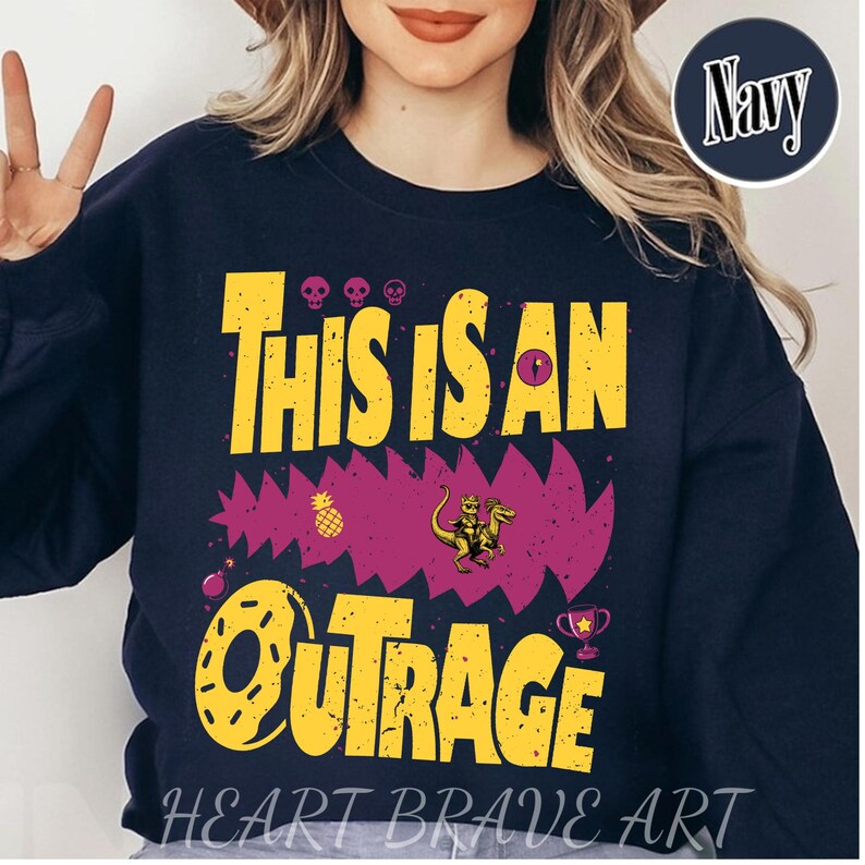 Puede incluir: Sudadera azul marino con el texto amarillo "THIS IS AN OUTRAGE", con un gr&aacute;fico de un dinosaurio montando un dinosaurio. Incluye un dise&ntilde;o de pi&ntilde;a y trofeo. Las palabras "HEART BRAVE ART" est&aacute;n en la parte inferior.