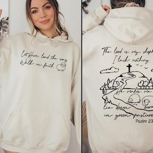 May include: A cream-coloured hoodie with a black graphic print of a sheep and the text "Let Him lead the way. Walk in faith." on the front. The back of the hoodie has a black graphic print of a hill with a cross on top and the text "The Lord is my shepherd: I lack nothing. He makes me lie down in green pastures. Psalm 23:1-2."