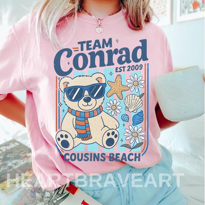May include: Pink t-shirt with a graphic featuring a bear wearing sunglasses and a scarf, surrounded by seashells and flowers. Text reads "TEAM Conrad EST 2009" and "COUSINS BEACH". The shirt is from HEARTBRAVEART.