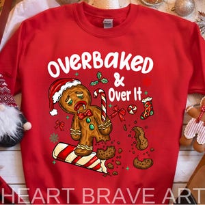 May include: Red sweatshirt with the text "OVERBAKED & Over It" and a gingerbread man wearing a Santa hat. The gingerbread man is slumped over a candy cane, with broken cookie pieces around it. Christmas-themed design.