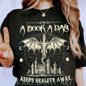 Pode incluir: Camiseta preta com um design gráfico branco que apresenta um dragão, um castelo e o texto "A Book A Day Keeps Reality Away".