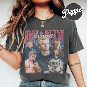 May include: A charcoal gray short-sleeve t-shirt featuring a vintage-style graphic print with the text DEANDI and Laurentis. The design includes multiple photographic portraits of a person, including one wearing a hockey jersey with the text BRIAR.