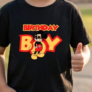 May include: A black short-sleeved t-shirt featuring a graphic of Mickey Mouse wearing sunglasses. The text "BIRTHDAY BOY" is printed in large yellow and red letters across the chest, with the character positioned in the center of the design.