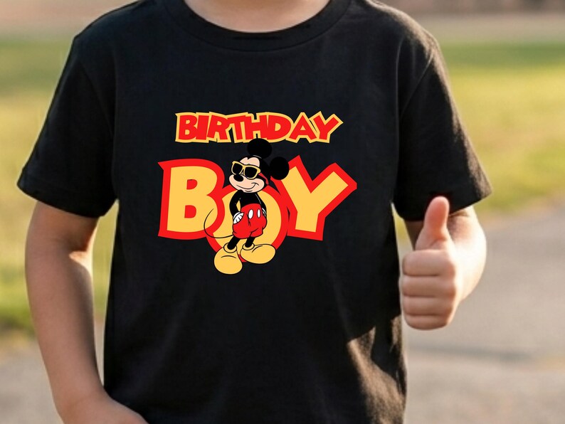 May include: A black short-sleeved t-shirt featuring a graphic of Mickey Mouse wearing sunglasses. The text "BIRTHDAY BOY" is printed in large yellow and red letters across the chest, with the character positioned in the center of the design.