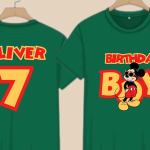 May include: Two dark green t-shirts featuring yellow and red text and graphics. One shirt displays the name OLIVER above the number 7. The other shirt features the text BIRTHDAY BOY with a graphic of Mickey Mouse wearing sunglasses.