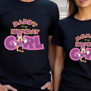 May include: Two black short-sleeved t-shirts featuring a graphic of Minnie Mouse. One shirt displays the text DADDY OF THE BIRTHDAY GIRL in yellow and purple, while the other shirt displays the text MOMMY OF THE BIRTHDAY GIRL in the same colors.