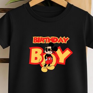 May include: A black short-sleeved t-shirt featuring a graphic of Mickey Mouse wearing sunglasses. The text "BIRTHDAY BOY" is printed in large yellow letters with a red outline above and behind the character, who wears red shorts and yellow shoes.