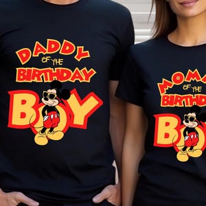 May include: Two black short-sleeved t-shirts featuring a graphic of Mickey Mouse wearing sunglasses. One shirt displays the text "DADDY OF THE BIRTHDAY BOY" in yellow and red, while the other shirt displays "MOMMY OF THE BIRTHDAY BOY" in the same colors.
