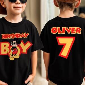 May include: A black t-shirt featuring a graphic of Mickey Mouse wearing sunglasses with the text BIRTHDAY BOY on the front. The back of the shirt displays the name OLIVER in red letters above a large yellow number 7 with a red outline.