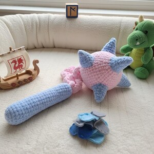 May include: A crocheted mace toy featuring a light pink spherical head with light blue spikes and a light blue cylindrical handle. A small pile of blue and gray felt pieces sits nearby, alongside a wooden toy ship and a green plush dragon.