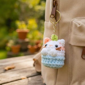 Puede incluir: Un llavero de ganchillo con forma de gato blanco con orejas y bigotes de color melocotón, sentado dentro de una cesta texturizada de color azul claro. El gato tiene ojos de cuentas negras y un pequeño broche verde en la cabeza.