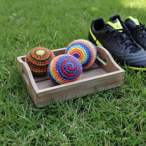 May include: Three spherical, crocheted yarn balls with colorful patterns resting in a small, rectangular wooden tray. One ball features a dark green, orange, and yellow design, while the other two display vibrant, multi-colored rainbow spirals.
