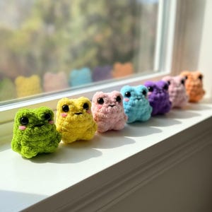 Peut inclure: Une rangée de sept petites grenouilles en crochet, douces et rembourrées, de différentes couleurs : vert, jaune, rose clair, bleu clair, violet, vieux rose et orange. Chaque grenouille a de grands yeux noirs et des joues roses.