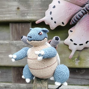 May include: A handmade crochet Blastoise plushie, featuring a blue and tan color scheme. The plushie has a round body, blue limbs, and a detailed shell. It is perched on a wooden post, with a decorative butterfly in the background.