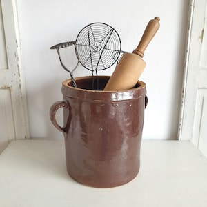 Peut inclure: Un pot en céramique marron avec deux poignées, contenant un rouleau à pâtisserie en bois et une passoire en fil de fer. Le pot a une finition émaillée et un aspect rustique, peut-être vintage. Les articles sont présentés sur une surface blanche.